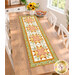 The completed Fancy Star Table Runner, colored in warm fabrics from the Sunflower Whispers collection by Riley Blake Designs, staged on a wood table with coordinating décor, white chairs, speckled with sunlight.