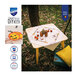 Lifestyle shot of the completed Halloween tablecloth staged on a small table outdoors among fallen leaves, staged with coordinating décor and lantern fruit; a close up on the jack-o'-lantern is also shown.