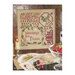 Finished sampler-style cross stitch pattern in a wooden frame placed on a table with doilies, candles, ornaments, and other holiday motifs.