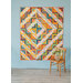 Colorful geometric patchwork quilt hanging on a blue wall above a wooden chair with flowers.