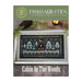 Front cover of the Cabin in the Woods cross stitch booklet with the finished pattern in a frame on a wooden table.