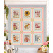 The completed Seed Packets quilt, colored in cream, green, pastel blue, and pink fabrics from the My Secret Garden collection by Moda Fabrics, hung on a rustic porch wall and staged with coordinating flowers and gardening tools.