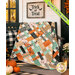 The completed Picnic Quilt Kit, colored in gray, mint green, white, pink, and orange fabrics from the Frank and Friends collection by Riley Blake Designs, artfully draped over a black porch bench beside pumpkins and a sign that says "Trick or Treat." A neon green banner in the top left corner reads "Glows in the dark."