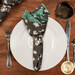 A top down shot of the napkin with the glow in the dark gray ghost fabric facing out, staged on a coordinating table setting on a warm wood table.