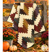 The completed Ridiculously Easy Jelly Roll Quilt, colored in warm, rustic prints from the Bountiful Blessings collection by Henry Glass Fabrics and artfully draped over a wooden fence, staged with rich coordinating florals and pumpkins.