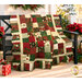 The completed Holiday Charm Quilt, colored in red, green, cream, and gold metallic fabrics from the Poinsettias at Christmas collection by Jennifer Bosworth for Riley Blake Designs, artfully draped over a black bench on a porch and staged with coordinating flowers and décor.