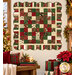 The completed Holiday Charm Quilt, colored in red, green, cream, and gold metallic fabrics from the Poinsettias at Christmas collection by Jennifer Bosworth for Riley Blake Designs, hung on a cream living room wall and staged with coordinating flowers and décor.