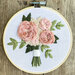 Round embroidery hoop displaying a hand-stitched bouquet of pink roses and green leaves on light fabric.