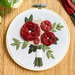 Round embroidery hoop displaying a hand-stitched bouquet of red roses and green leaves on light fabric.