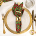 A top down shot of the napkin with the red and green fabric facing out, staged on a coordinating gold table setting on a white marble table.