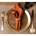 A top down shot of the napkin with the orange paisley fabric facing out, staged on a coordinating gold table setting on a white table.