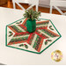 The completed 60 Degree Diamond Table Topper colored in cream, red, and green fabrics with gold metallic accents from the Adorn collection by Andover Fabrics, staged on a white table with a frosted green jar holding greenery in the center.