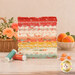 A stack of the fabrics included in the Apricot Candy collection, with a basket and flowers and apricots in a bowl and candies and thread.