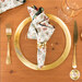 A top down shot of the napkin with the cream fabric facing out, staged on a coordinating gold table setting on a white table.