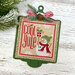 Green wooden ornament display with a mounted cross stitch design reading "Warm Wishes," with a snowman, gift, snowflakes, evergreen sprigs, pinecone, and red-and-white striped bow.
