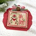 A Christmas cross stitch ornament with a wide-eyed reindeer wearing a green scarf, tiny ornaments hanging from its antlers, and a sprig of holly.