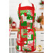 The completed Charm Squares Apron, colored in bright red, green, and white fabrics from the Snow Time collection by Moda Fabrics, staged on a mannequin beside coordinating décor and a Christmas tree.