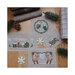 Photo of the finished cross stitch patterns laid flat on a wooden surface next to scattered pine sprigs and snowflakes.
