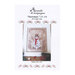 Front cover of the cross stitch pattern with the finished pattern placed in a white frame lying with pine boughs on a wooden table.