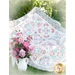 A colorful quilt with a geometric pattern in pastel shades is draped over a grassy area. Next to the quilt, there is a white vase filled with pink and purple flowers.