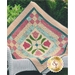 A quilt with a patchwork design featuring pink tulips and green leaves at the center, surrounded by a checked and solid fabric border in shades of pink, blue, and cream. The quilt is draped over a chair, with a logo for Shabby Fabrics in the corner.