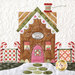 A whimsical fabric block depicting a quilt shop designed like a gingerbread house, featuring a brown facade, colorful windows, and decorative candy cane fences. The sign reads Ginger's Quilt Shoppe, and a spool of thread sits atop the roof.