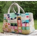 A colorful quilted tote bag featuring a patchwork design of houses in various colors and patterns, with sturdy circular handles. The bag is placed on a rustic wooden surface against a green outdoor background.