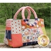 A colorful quilted handbag with floral and geometric patterns, featuring two rounded handles, placed on a wooden surface with a green outdoor background.