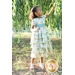 A young girl with curly hair stands outdoors, reaching for a branch of a tree. She wears a pastel-colored, tiered dress with floral patterns and a blue ribbon.