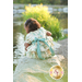 A young girl in a floral dress sits on a rock by a gentle body of water, her back turned towards the camera. Lush greenery surrounds the scene.