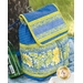 A blue and yellow floral patterned backpack sits against a tree. The bag features a folded flap and decorative stripes, with vibrant flowers and polka dots on its surface. A green bottle is partially visible next to the backpack.