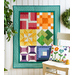 A colorful quilt hanging on a wall, featuring various patterns and shapes in bright colors, with a small decorative pillow on a chair nearby. A green plant is also visible in the background.