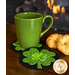 A green mug sits on a black circular coaster adorned with a bright green shamrock. Next to the mug is a flaky croissant. In the background, a fireplace is softly flickering.