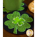 A green mug sits on a round black coaster featuring a large, green shamrock design, surrounded by a croissant on a wooden table.