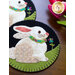 Two circular wool Mug Rugs featuring white rabbits sitting on green grass. Each rabbit has a colorful collar and is surrounded by floral decorations. The background is black, and the ornaments have a stitched border.