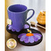 A purple mug sits on a decorative black coaster with a flower design. The coaster features petals in shades of purple and an orange center, with a small bee. Nearby, there is a honey jar and a honey dipper.