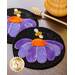 Two circular wool coasters featuring a purple flower design with an orange center and a small bee. The background is black, and the coasters are placed on a wooden surface alongside a honey dipper and a honey jar.