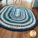 An oval braided rug in shades of blue, teal, and cream is placed on a wooden floor. The rug has a textured pattern, and wooden furniture legs are visible in the background.