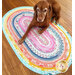 A brown dog lies on a colorful oval fabric rug with a floral pattern. The rug features vibrant hues of pink, blue, and orange. The background shows a wooden floor.