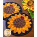 Two round coasters designed as sunflowers, featuring yellow petals, a brown center with seeds, and a small ladybug. The background is dark, complementing the bright colors of the flowers. A sunflower decoration is visible in the background, along with a logo for Shabby Fabrics.