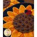 A close-up of a decorative wool sunflower mug rug coaster with vibrant yellow petals and a textured brown center, featuring a small ladybug on top. The background includes additional sunflowers, creating a warm, colorful display.