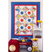 A colorful quilt featuring a geometric design with flower appliqués in red and blue, hanging on a wall. Next to it, a white shelf holds a vase of yellow flowers, books, and a small clock. A red wagon with a blanket and items is positioned in front of the shelf.