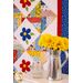 A vibrant quilt featuring colorful patchwork squares and floral patterns is displayed on a wall, alongside a silver metal pitcher filled with bright yellow flowers. Stacked glass jars sit next to the pitcher on a white surface.