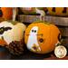 A collection of decorative felt pumpkins in various colors, including orange and cream. One orange pumpkin features a white ghost and a black spider as embellishments. Small pinecones are nearby, adding to the autumn theme.