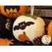 A decorative white felt pumpkin featuring a black bat and an orange pumpkin design on its surface, surrounded by other colorful pumpkins and autumn-themed decorations.