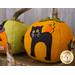 A decorative felt pumpkin in orange with a black cat design on the front, featuring a green top and an orange detail on the cat's tail. Another pumpkin in green with a playful face is partially visible in the background.