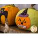 Two decorative pumpkins made of felt sit on a wooden surface. One pumpkin is orange with a black cat, and the other is green with a jack-o-lantern and purple witch's hat.