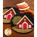 Two round coasters featuring a decorative design of a small red house with a bell on the roof, surrounded by trees and flowers on a black background. Slices of apple and cinnamon sticks are placed nearby on a wooden surface.