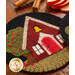 A round, wool felt coaster featuring a colorful design of a small house with a red door and a bell, surrounded by a tree with red apples. The background is black, and the coaster has a stitched border. In the background, there are cinnamon sticks and apple slices on a wooden table.