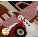 A close-up view of a fabric design featuring a red truck with a snowman beside it. The snowman has a carrot nose, buttons, and a red scarf. The background includes various textures and colors.